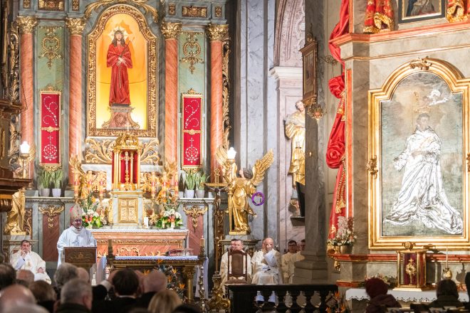 Uroczysta msza święta w barokowym kościele. Biskup w białych szatach liturgicznych przemawia z ambony, a kapłani siedzą wokół ołtarza. W tle bogato zdobiona ściana z obrazami i figurami świętych