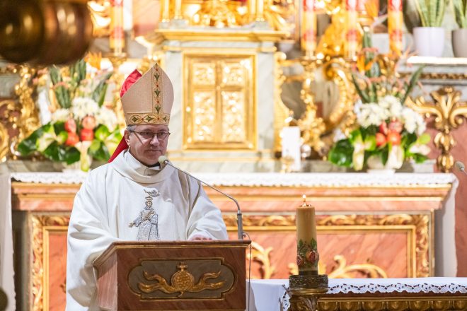 Zbliżenie na biskupa w ceremonialnym stroju, przemawiającego zza ambony. Jego mitra ozdobiona jest złotymi haftami, a za nim widoczny jest ołtarz udekorowany świeżymi kwiatami i złoceniami