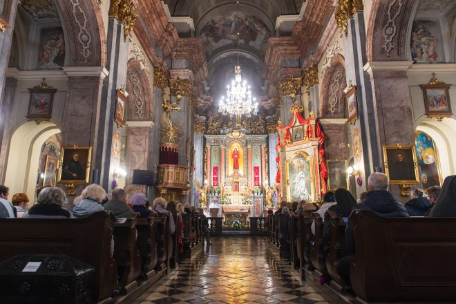 Wnętrze barokowego kościoła wypełnione wiernymi. Na ołtarzu bogato zdobione elementy liturgiczne, złocone figury aniołów i duży obraz Chrystusa. Po prawej stronie wizerunek świętego w ozdobnej ramie