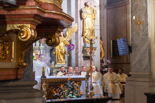 Kapłan stojący przy ołtarzu okadza święte miejsce, unosząc kadzielnicę. W tle duchowni w złotych ornatach siedzą na ławach, wsłuchani w liturgię. Na ekranie wyświetlony jest tekst modlitwy