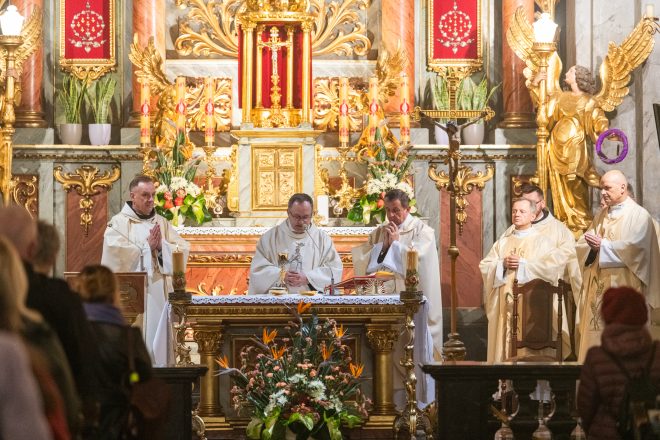 Celebracja Eucharystii przy bogato zdobionym ołtarzu. Kapłani w skupieniu odprawiają mszę, jeden z nich podnosi kielich mszalny. W tle złocone figury aniołów oraz ozdobne świece