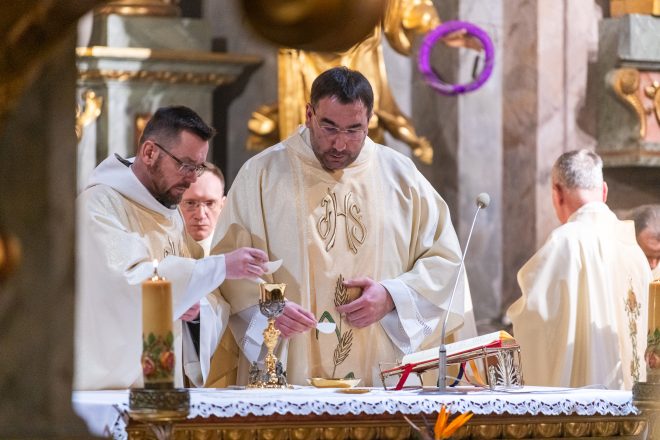 Moment konsekracji – dwóch kapłanów przygotowuje Hostię i wino na ołtarzu. Jeden z nich ostrożnie kładzie komunikant na kielich, podczas gdy drugi trzyma patenę. W tle złote dekoracje świątyni