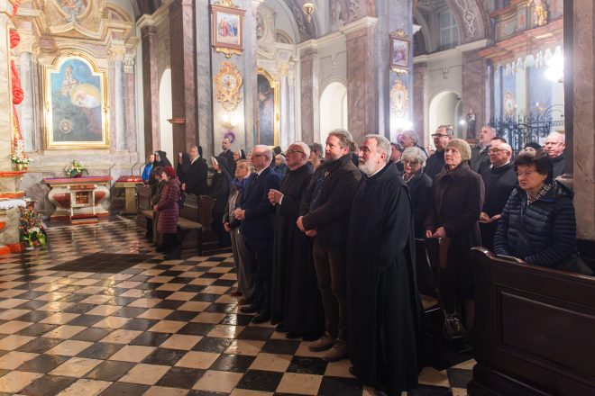 Wierni zgromadzeni w kościele stoją w modlitewnym skupieniu. Wśród nich duchowni, zakonnice oraz świeccy, odziani w ciepłe ubrania. W tle bogato zdobione wnętrze świątyni z obrazami i rzeźbami