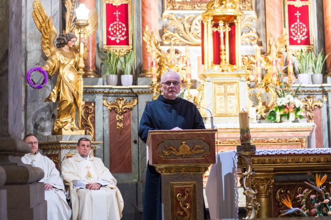 Brat zakonny w mitrze, stojący przy ambonie, wygłasza kazanie. W tle złocony ołtarz z figurą anioła trzymającego świecę i fioletowy wieniec liturgiczny.