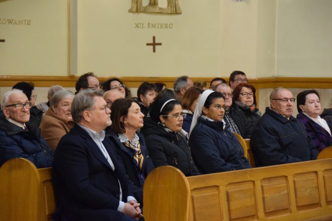 Wnętrze kościoła z liczną grupą wiernych stojących w ławkach. Wierni są skupieni, część ma złożone dłonie, a w tle widać organy oraz dekoracje ścienne przedstawiające stacje drogi krzyżowej