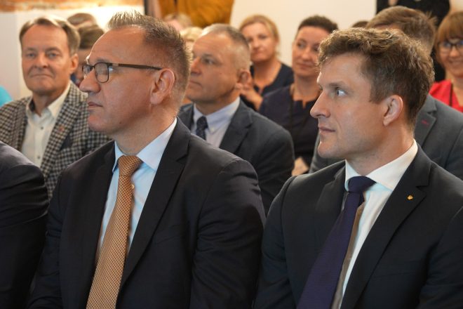 Dwóch elegancko ubranych mężczyzn siedzi w pierwszym rzędzie podczas oficjalnego wydarzenia. Mają na sobie garnitury i krawaty, a wokół nich widać zgromadzoną publiczność, uważnie śledzącą przebieg spotkania