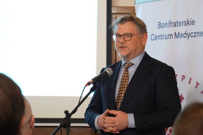 Mężczyzna w garniturze i okularach przemawia do mikrofonu, stojąc przed banerem Bonifraterskiego Centrum Medycznego. W tle widać ekran z prezentacją