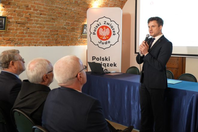 Mężczyzna w granatowym garniturze przemawia do publiczności, trzymając mikrofon. Za nim znajduje się baner Polskiego Związku Judo, a w tle widać zabytkowe ceglane sklepienie