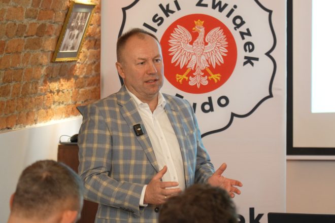 Mężczyzna w jasnej marynarce w kratę gestykuluje podczas przemowy. Za nim widoczny jest baner Polskiego Związku Judo oraz ekran prezentacyjny