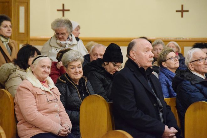 Starsze osoby siedzące w drewnianych ławkach wewnątrz kościoła, uczestniczące w nabożeństwie. Część z nich ma złożone ręce, inne patrzą skupione na ołtarz. W tle widać inne osoby, niektóre stojące, a na ścianach umieszczone są symbole krzyża i elementy dekoracyjne kościoła