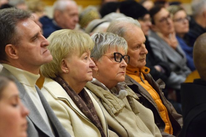 Wierni siedzący w ławkach kościoła, skupieni i zasłuchani. Wśród nich starsze osoby w eleganckich płaszczach i szalach, a w tle kolejne rzędy parafian uczestniczących w nabożeństwie