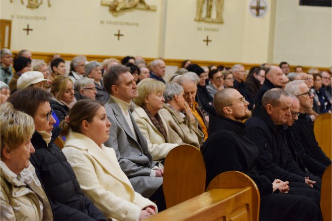 Zbliżenie na wiernych siedzących w ławkach kościoła, ubranych w eleganckie płaszcze. Wśród nich zakonnik w czarnym habicie oraz starsi mężczyźni w ciemnych marynarkach