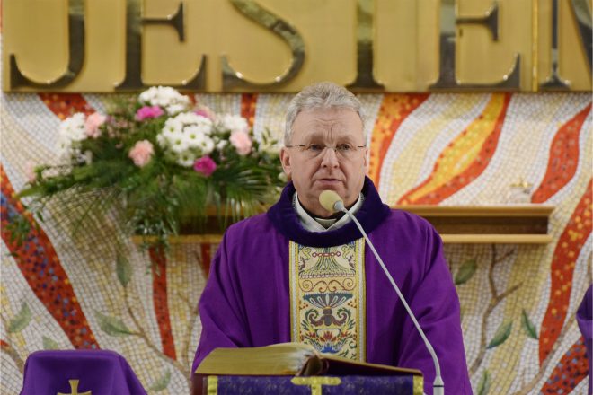 Kapłan w fioletowym ornacie stoi przy ambonie na tle mozaiki płomieni i złotego napisu „JESTEM”. Przed nim otwarta księga liturgiczna, a wokół kwiatowe dekoracje