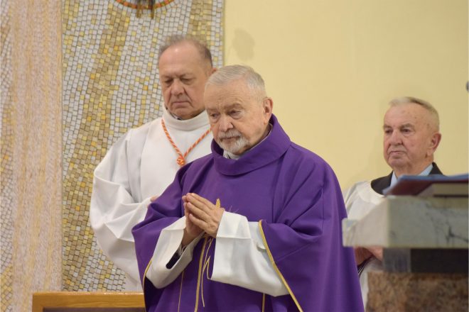 Starszy duchowny w fioletowym ornacie składa dłonie w geście modlitwy. Za nim inni kapłani w białych albach uczestniczą w uroczystości liturgicznej