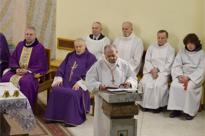 Kapłan w białej albie i fioletowym ornacie czyta tekst liturgiczny z ambony, za nim siedzi kilku duchownych i ministrantów ubranych w białe alby. Uroczysta atmosfera nabożeństwa