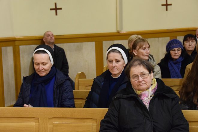 Zbliżenie na grupę starszych osób siedzących w ławkach kościelnych. Wśród nich widać dwie zakonnice w ciemnych habitach oraz kilku mężczyzn i kobiet w zimowych ubraniach