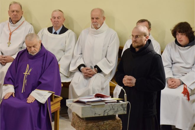 Młody zakonnik w czarnym habicie stoi przy ambonie, z rękami złożonymi do modlitwy. Obok niego duchowni i ministranci w białych szatach uczestniczą w liturgii