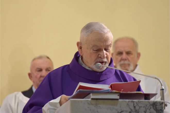 Starszy kapłan w fioletowym ornacie wygłasza kazanie, czytając z otwartej księgi liturgicznej. Za nim siedzą inni duchowni, skupieni na przebiegu mszy