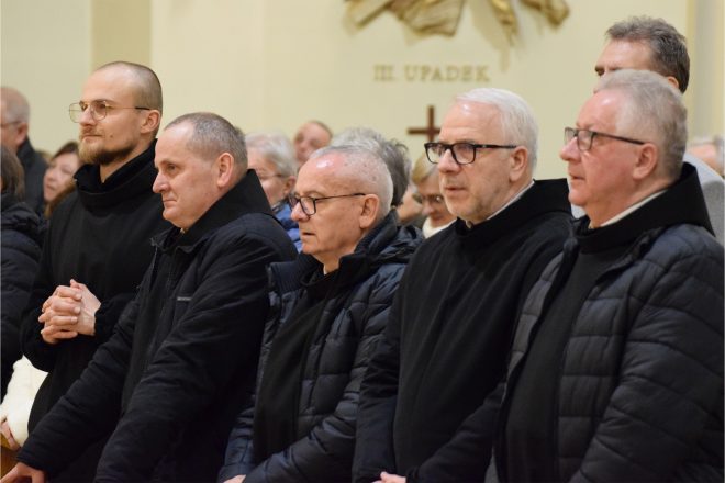 Grupa zakonników i duchownych w ciemnych habitach i płaszczach, stojących w ławkach kościelnych. Niektórzy z uśmiechem na twarzy, inni w skupieniu.