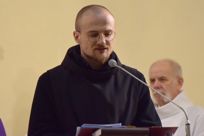 Młody zakonnik w czarnym habicie stoi przy ambonie i czyta tekst na głos, koncentrując się na słowach. Jego twarz oświetlona jest ciepłym światłem, a w tle widać starszego kapłana w białej albie, wsłuchanego w przemówienie