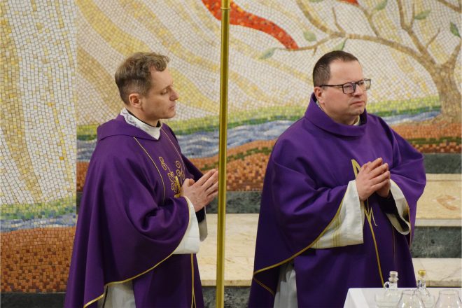 Dwóch kapłanów w fioletowych szatach liturgicznych stoi w skupieniu przy ołtarzu. Jeden z nich ma złożone dłonie w geście modlitwy, drugi patrzy w stronę celebransa. W tle mozaika przedstawiająca drzewo i płomienie