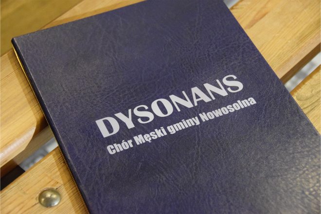 Zbliżenie na śpiewnik chóru „Dysonans”, oprawiony w granatową okładkę ze srebrnym napisem: „DYSONANS – Chór Męski gminy Nowosolna”. Śpiewnik leży na drewnianej ławie