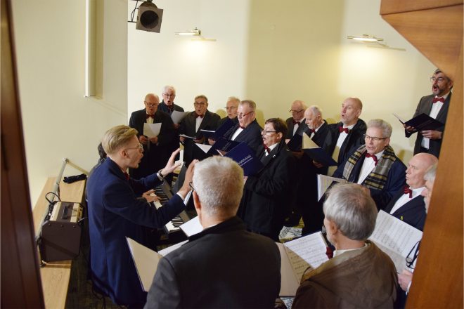 Chór męski wykonuje pieśni na chórze kościelnym pod dyrygenturą młodego kapelmistrza. Śpiewacy, ubrani w eleganckie garnitury i muszki, trzymają śpiewniki, z których odczytują nuty. W tle jasne ściany kościoła i drewniana bariera chóru