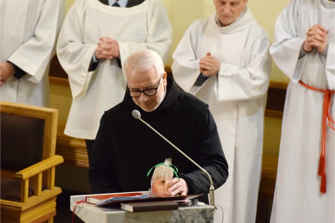 Brat zakonny w czarnej szacie zakonnej wygłasza kazanie z ambony, trzymając w ręce jabłko zapakowane w celofan z zieloną wstążką. W tle biali ministranci z dłońmi splecionymi w geście modlitwy.