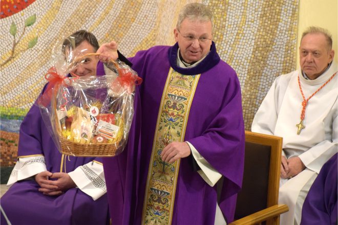 Ksiądz w bogato zdobionej fioletowej szacie liturgicznej prezentuje kosz upominkowy owinięty w przezroczystą folię i ozdobiony czerwoną wstążką. Obok niego duchowny w podobnym stroju z uśmiechem spogląda na prezent