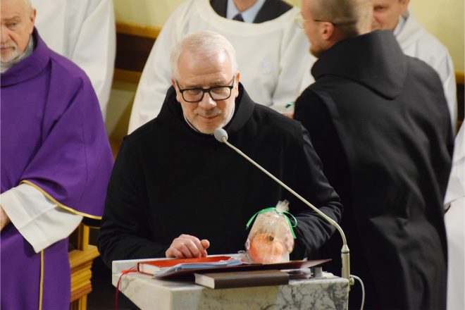 Przemówienie zakonnika w czarnej szacie przy ambonie. Na pierwszym planie owoc w ozdobnym opakowaniu, który trzyma w dłoni. W tle księża ubrani w liturgiczne szaty w kolorze fioletowym oraz białe alby ministrantów