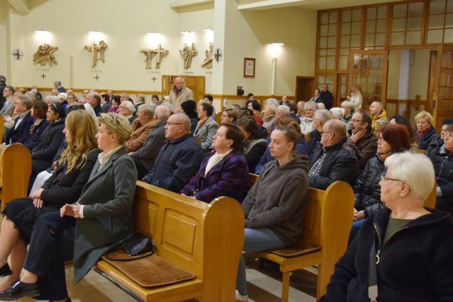 Wnętrze kościoła z liczną grupą wiernych stojących w ławkach. Wierni są skupieni, część ma złożone dłonie, a w tle widać organy oraz dekoracje ścienne przedstawiające stacje drogi krzyżowej