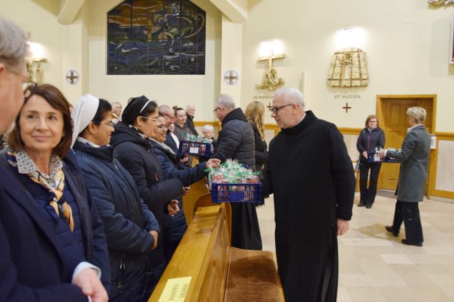 Bracia oraz wolontariusze rozdają symboliczne jabłka uczestnikom uroczystości. Wierni, w tym zakonnice w ciemnych habitach, odbierają owoce z wdzięcznością i uśmiechem. W tle rozświetlone wnętrze kościoła.