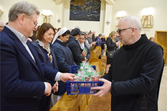 Mężczyzna w ciemnym habicie zakonny odbiera plastikowy koszyk z zapakowanymi jabłkami od elegancko ubranego mężczyzny. W tle widać siostry zakonne oraz innych uczestników uroczystości, czekających na swoją kolej, aby otrzymać symboliczne upominki