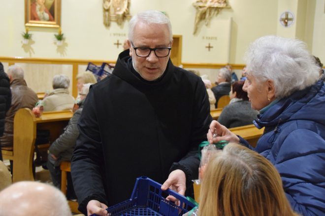 Zakonnik o siwych włosach i okularach rozdaje upominki starszym kobietom siedzącym w ławkach kościoła. Kobieta w niebieskiej kurtce sięga po owoc zapakowany w przezroczystą folię z zieloną kokardką, a w tle widać innych uczestników wydarzenia