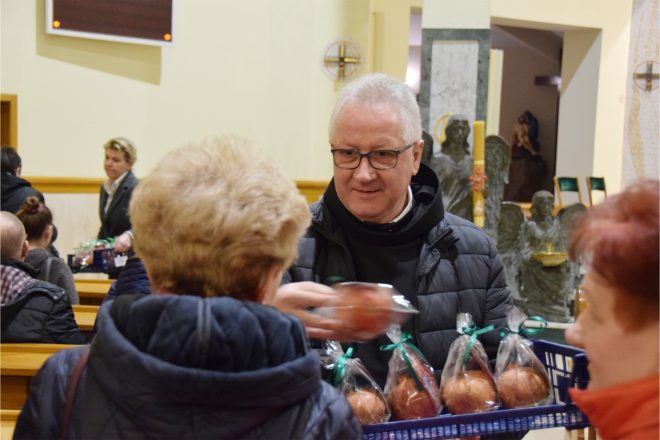 Zakonnik w czarnej kurtce uśmiecha się serdecznie, wręczając starszej kobiecie jabłko. Na pierwszym planie widać plastikowy koszyk wypełniony starannie zapakowanymi owocami, a w tle wnętrze kościoła i inne osoby biorące udział w uroczystości.