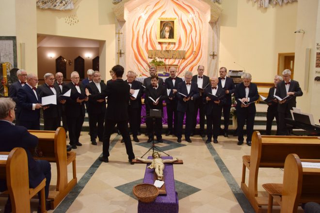 Męski chór w eleganckich garniturach i muszkach śpiewa pod dyrekcją dyrygenta, stojąc przed ołtarzem udekorowanym motywem płomieni i obrazem św. Faustyny. Na pierwszym planie leży krzyż z figurą Jezusa, a przed nim ustawiony jest koszyk na ofiary