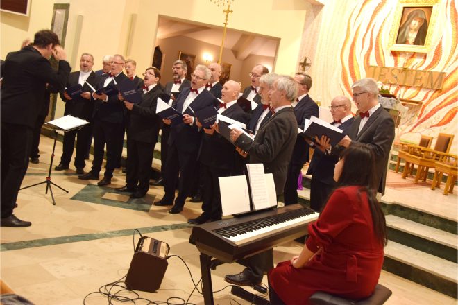 Chór męski występuje przed ołtarzem, śpiewając pod dyrekcją prowadzącego, który gestykuluje, nadając rytm występowi. Chórzyści trzymają czarne teczki z nutami, a w tle widać ołtarz ozdobiony ciepłym światłem oraz wizerunek świętej. Przed chórem stoi pianino elektryczne, przy którym siedzi akompaniująca artystka w czerwonej sukni