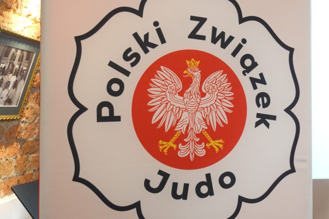 Zbliżenie na baner Polskiego Związku Judo, przedstawiający biało-czerwone logo z orłem w koronie na czerwonym tle. W tle ceglana ściana i zawieszone na niej zdjęcie