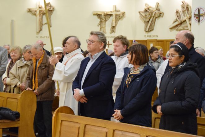 Uczestnicy nabożeństwa stojący w ławkach. Wśród nich widoczny mężczyzna w garniturze oraz kobieta w ciemnej kurtce i apaszce, a także zakonnica. Obok nich procesja liturgiczna z ministrantem niosącym świecę