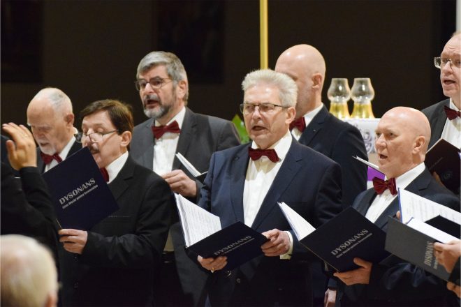 Grupa mężczyzn ubranych w eleganckie garnitury z muszkami śpiewa podczas uroczystości w kościele. W dłoniach trzymają nuty oznaczone nazwą chóru „Dysonans”. Jeden z mężczyzn, w okularach i z siwymi włosami, śpiewa z zaangażowaniem. W tle widać innych chórzystów oraz kościelne wnętrze z ołtarzem ozdobionym świecami i złoconymi detalami