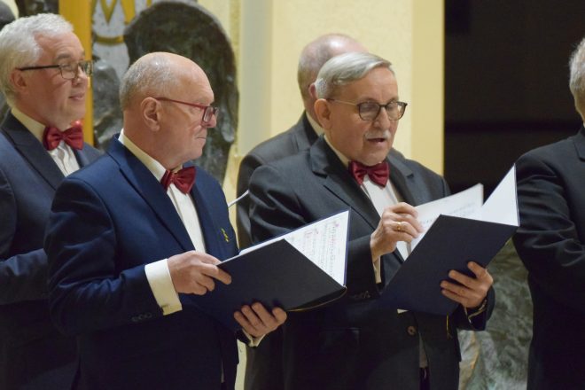 Dwaj starsi mężczyźni w garniturach i czerwonych muszkach śpiewają z nutami w rękach. Skupienie na ich twarzach podkreśla zaangażowanie w występ chóru