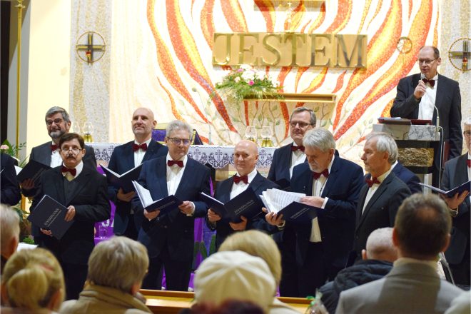 Kilku członków chóru trzymających nuty w rękach, śpiewających na tle bogato zdobionego ołtarza w kościele. W tle prowadzący wydarzenie mężczyzna przemawia do mikrofonu