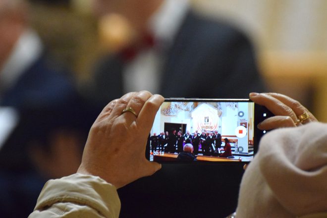 Zbliżenie na rękę osoby nagrywającej występ chóru na telefonie komórkowym. Na ekranie widać ujęcie chóru stojącego przed ołtarzem, w tle dekoracje kościelne