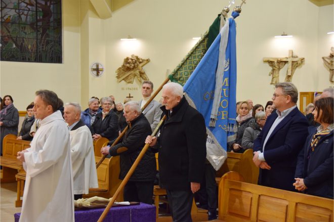 Procesja liturgiczna wewnątrz kościoła. Starszy mężczyzna niesie sztandar, a wokół niego zgromadzeni są inni uczestnicy uroczystości, w tym osoby w białych szatach liturgicznych