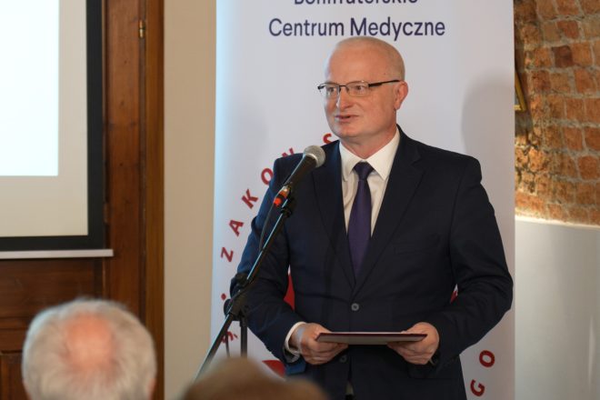 Mężczyzna w ciemnym garniturze i fioletowym krawacie przemawia do mikrofonu, trzymając w dłoniach dokument. W tle widoczny jest baner Bonifraterskiego Centrum Medycznego oraz ceglaste sklepienie sali