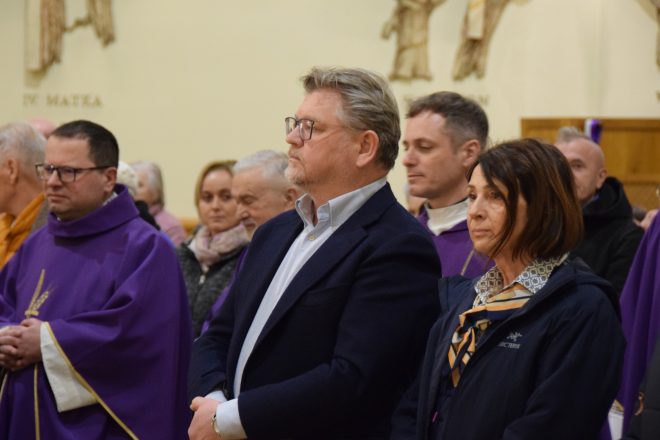 Zbliżenie na uczestników nabożeństwa, wśród nich mężczyzna w granatowym garniturze oraz kobieta w ciemnym ubraniu. W tle duchowni w fioletowych ornatach