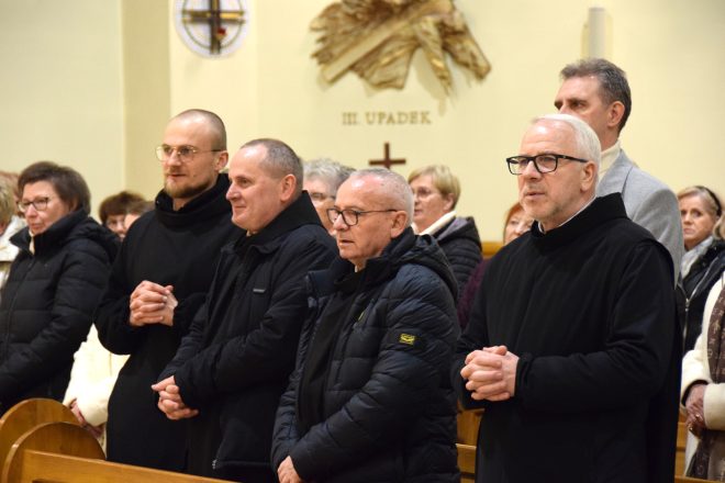 Grupa zakonników i duchownych w ciemnych habitach i płaszczach, stojących w ławkach kościelnych. Niektórzy z uśmiechem na twarzy, inni w skupieniu