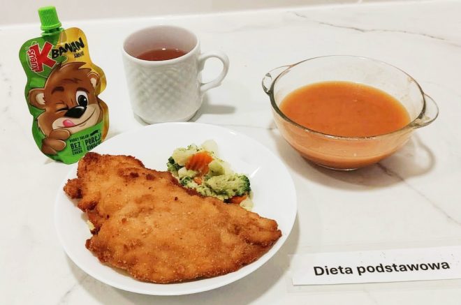 Na białym marmurowym blacie znajduje się zestaw obiadowy oznaczony etykietą „Dieta podstawowa”. Na dużym białym talerzu podano panierowany filet drobiowy oraz porcję gotowanych warzyw – brokułów, kalafiora i marchewki. Obok stoi miska z gorącą, gęstą zupą pomidorową. Dodatkiem jest owocowy mus w zielonym opakowaniu z wizerunkiem misia oraz filiżanka gorącego napoju, prawdopodobnie herbaty