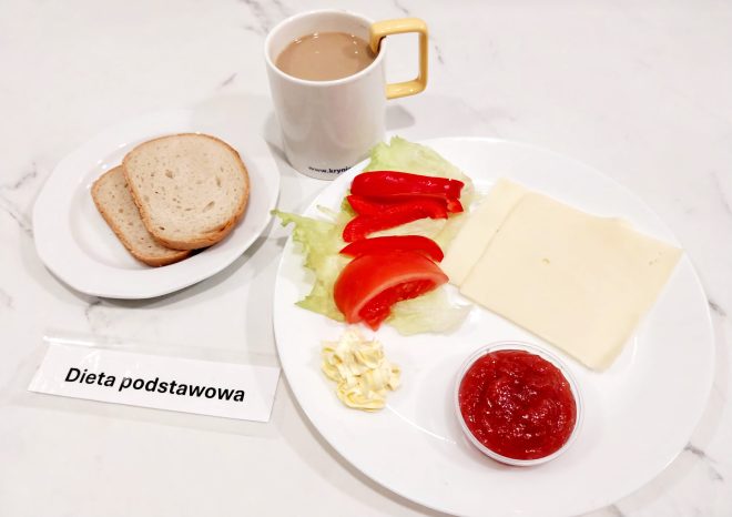 Zestaw śniadaniowy w ramach „Diety podstawowej” na jasnym blacie. Na głównym talerzu znajdują się dwa plastry białego sera żółtego, kilka kawałków sałaty lodowej, pokrojone plasterki czerwonej papryki i pomidora, porcja margaryny oraz mały pojemnik z konfiturą truskawkową lub przecierem owocowym. Obok na osobnym talerzyku – trzy kromki chleba pszennego. W tle biały kubek z napojem mlecznym lub kawą z mlekiem. Całość sfotografowana z góry. Na pierwszym planie widoczny podpis: „Dieta podstawowa”