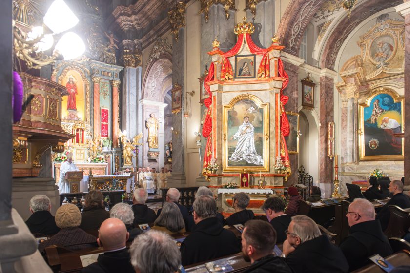 Fragment wnętrza kościoła z wiernymi siedzącymi w ławkach. Na pierwszym planie duży obraz świętego w złotej ramie, ozdobiony czerwoną tkaniną